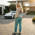 Model wearing a cream Need Money for Porsche hoodie from behind with rounded purple lettering and turquoise racing-style back graphic